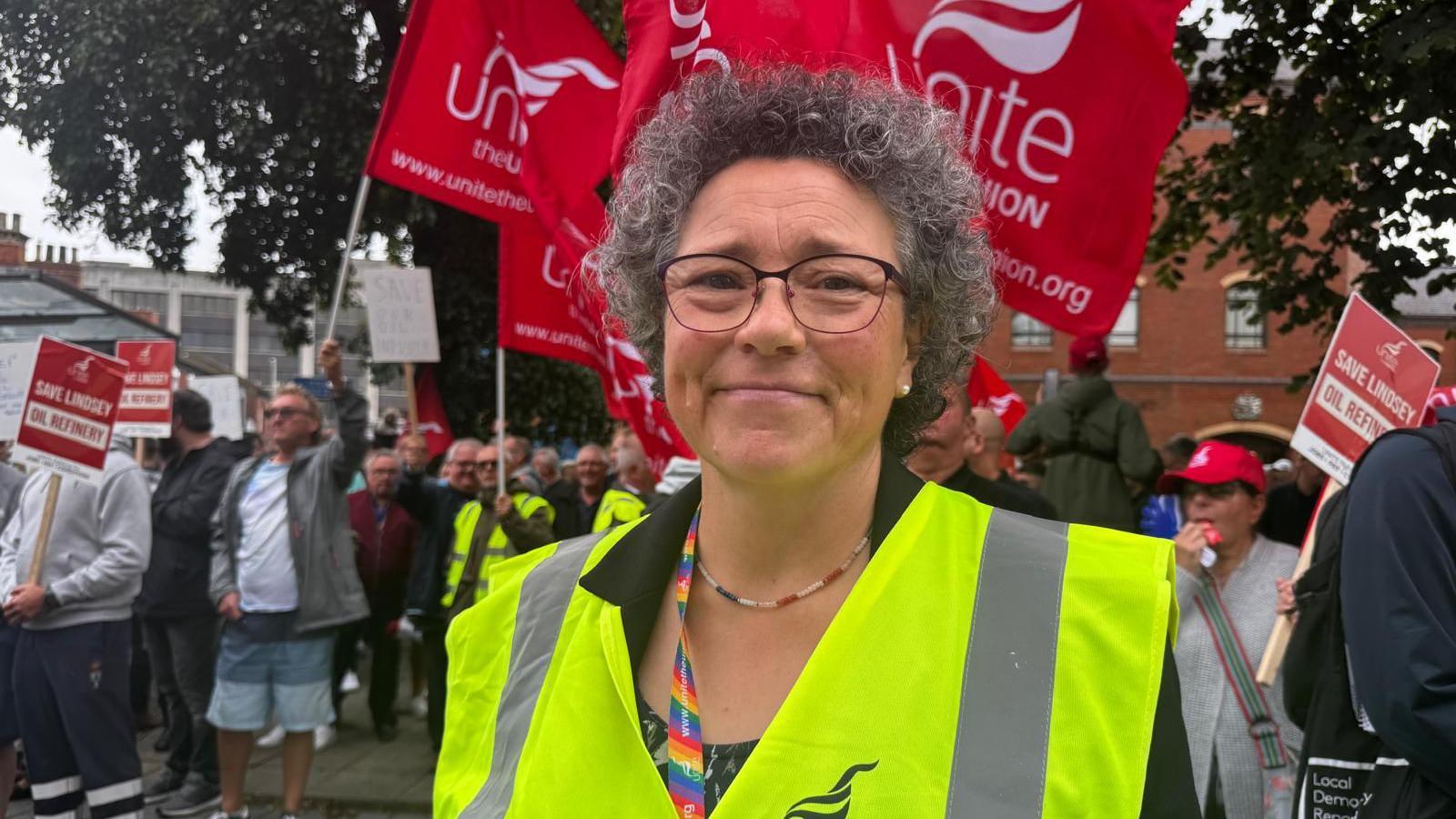 Woman with curly hair, glasses, fluorescent bib and dark top stood in front of a crowd of people with unite the Union flags.