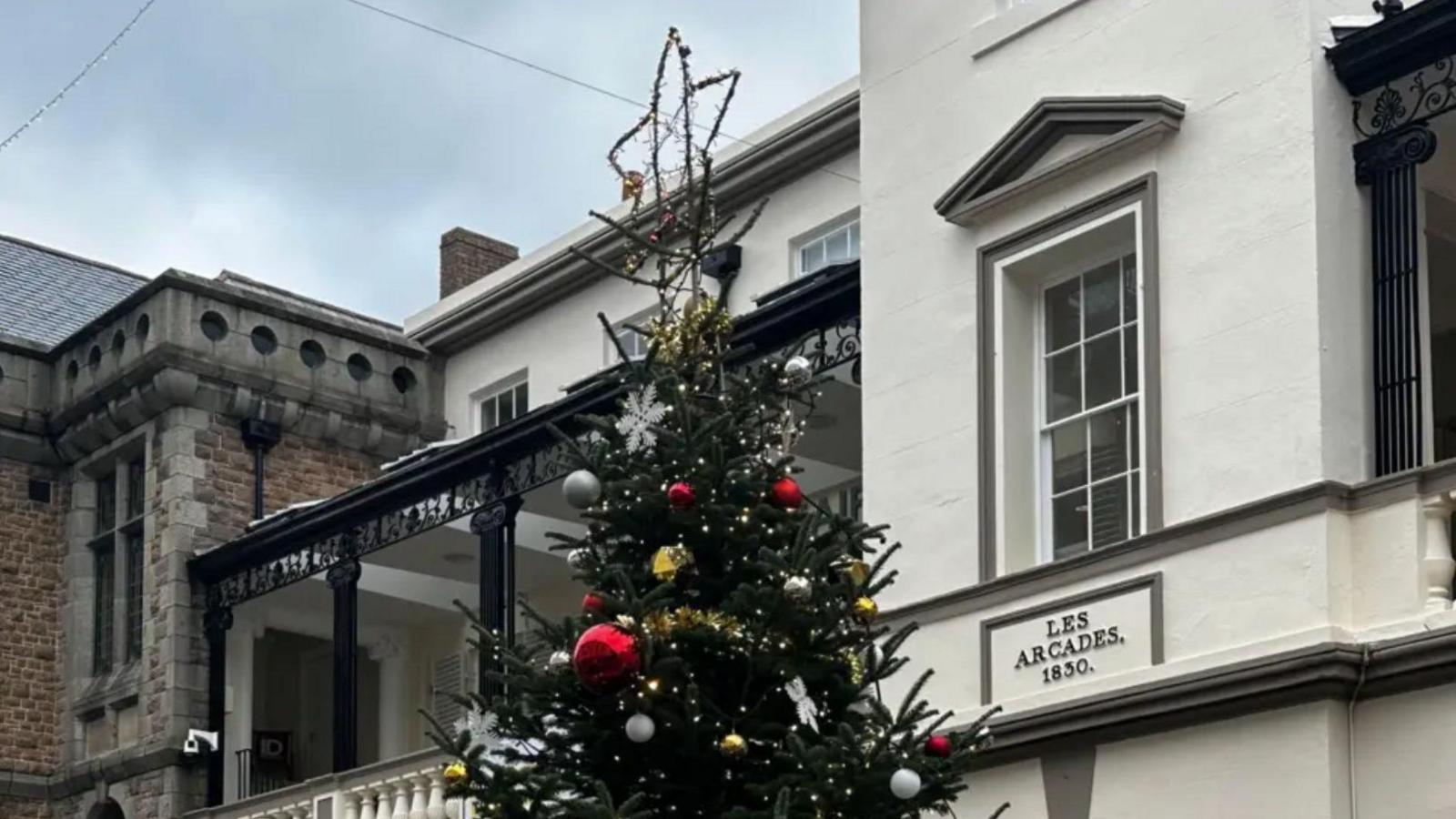 A Christmas tree pictured in the centre of St Peter port. You can only see the top of the Christmas tree with the star on it. There is a building which says Les arcades 1830 on it.