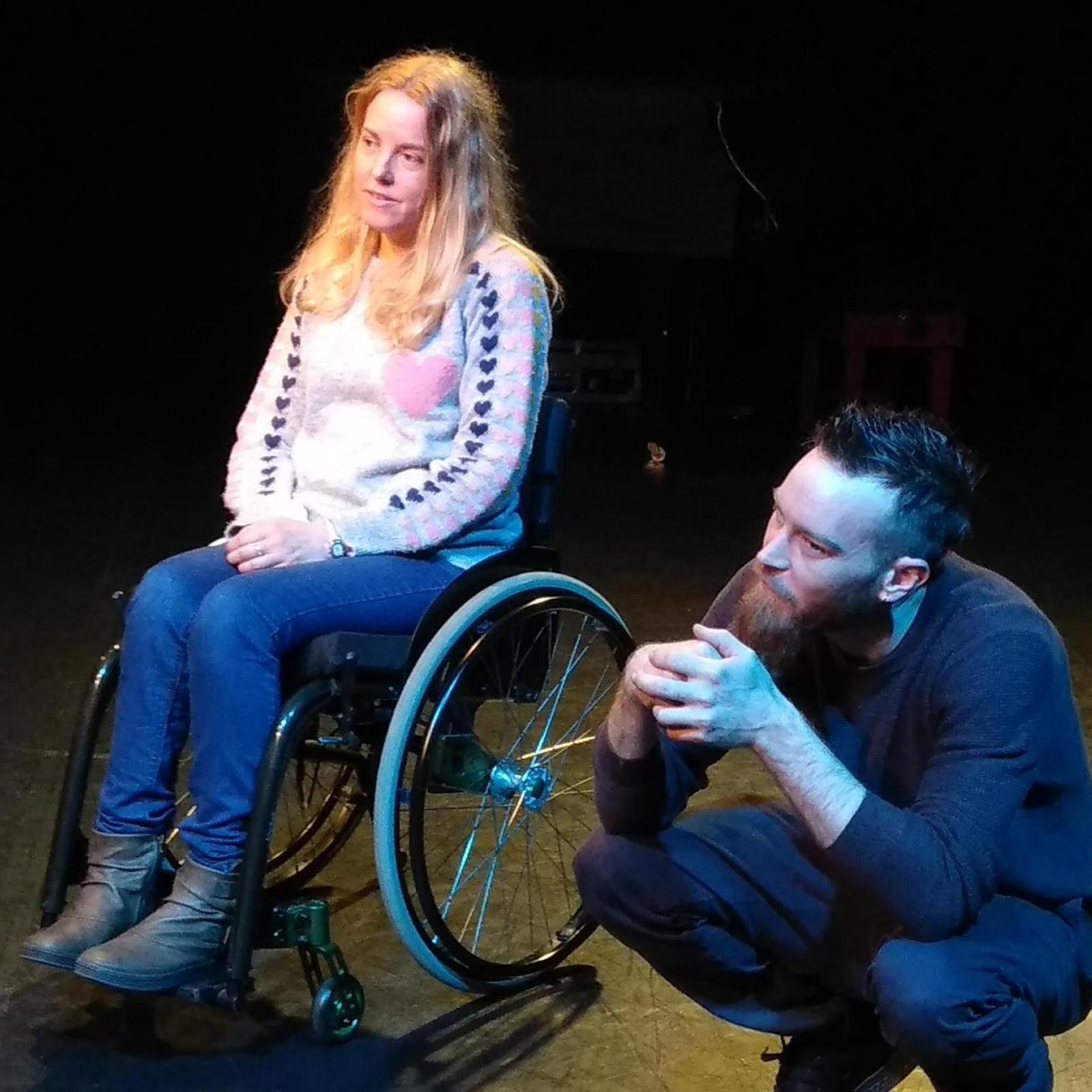 A blonde woman in a wheelchair next to a crouching man on an empty stage. She is wearing jeans and a pink jumper.