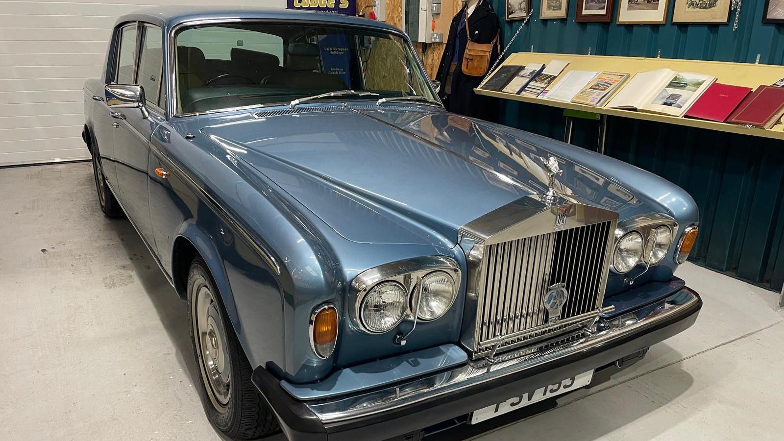 The blue Rolls Royce sits on display in the museum. An exhibition to its right with books and pictures can be seen as well as a dummy wearing an old bus driver uniform.