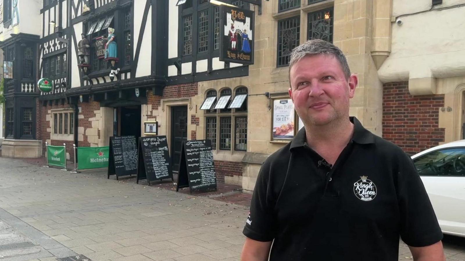 A man in a black polo shirt standing in front of an old pub.