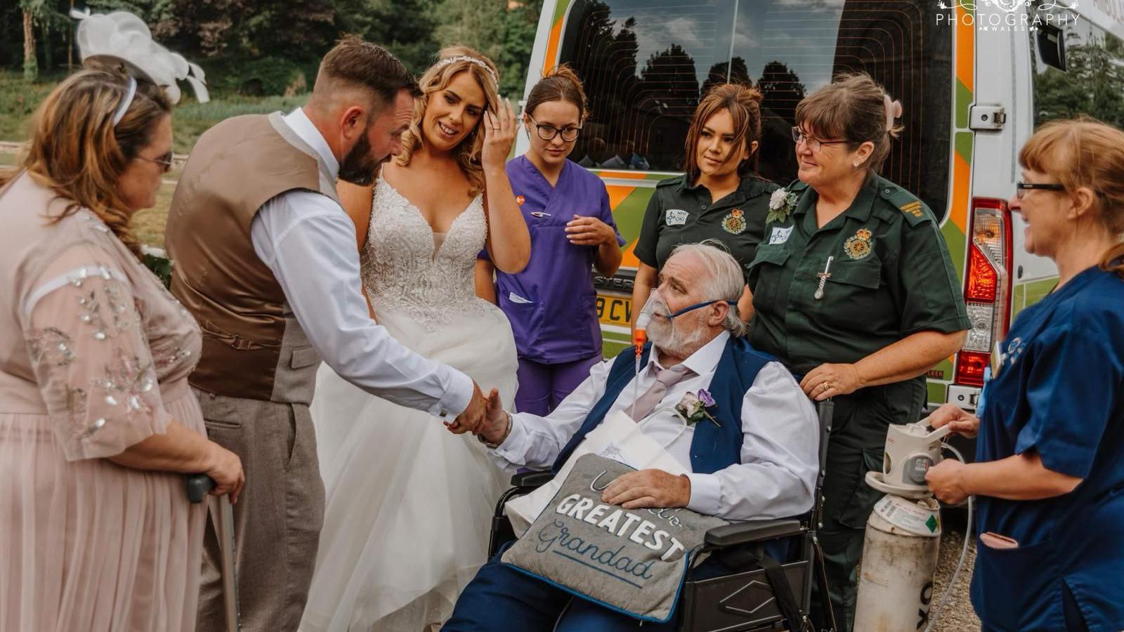 Wayne is in a wheelchair, with an oxygen mask, wearing a suit at his daughter's wedding. He is surrounded by two paramedics and two nurses, next to an ambulance. He is shaking the hand of his son-in-law, next to his daughter, who is wearing a wedding dress. His wife is also in the picture.