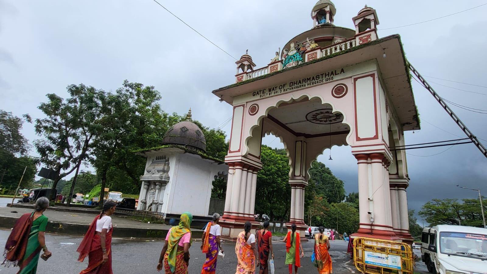 Women in colourful saris walk on a cloudy day towards an archway that's the entrance to the temple at Dharmasthala in Karnataka