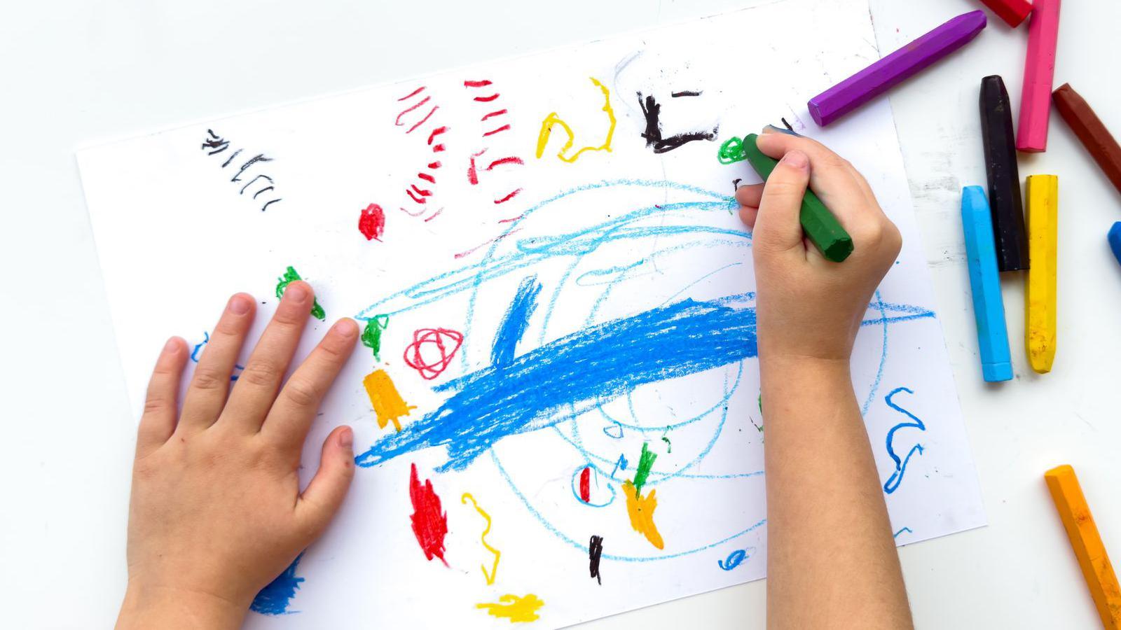 Childs hands drawing with colorful wax crayons on white paper, top view
