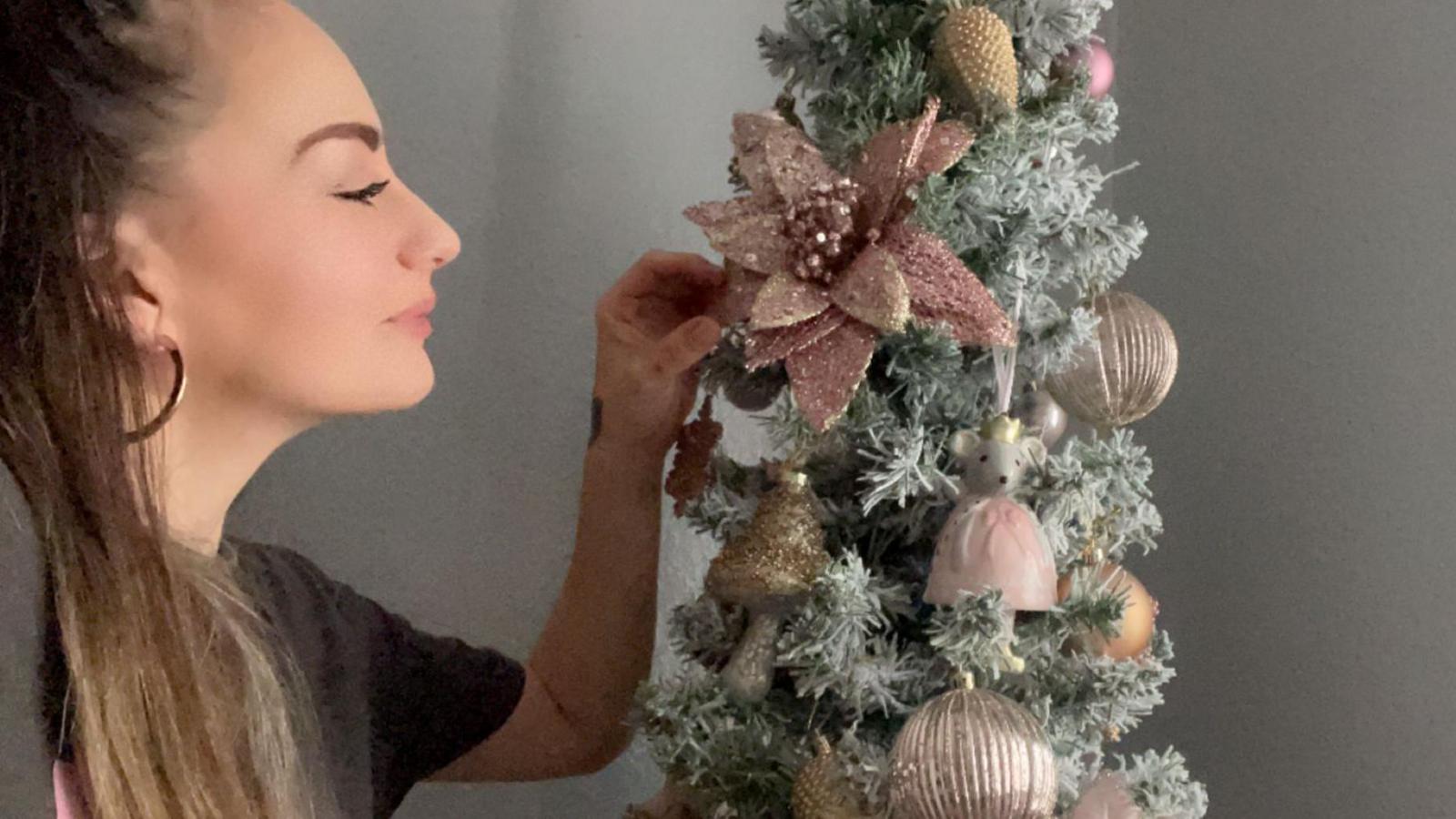 Sally-Anne Long, a woman with her hair tied back wearing a grey t-shirt, looks at a Christmas tree decorated with gold and red ornaments.