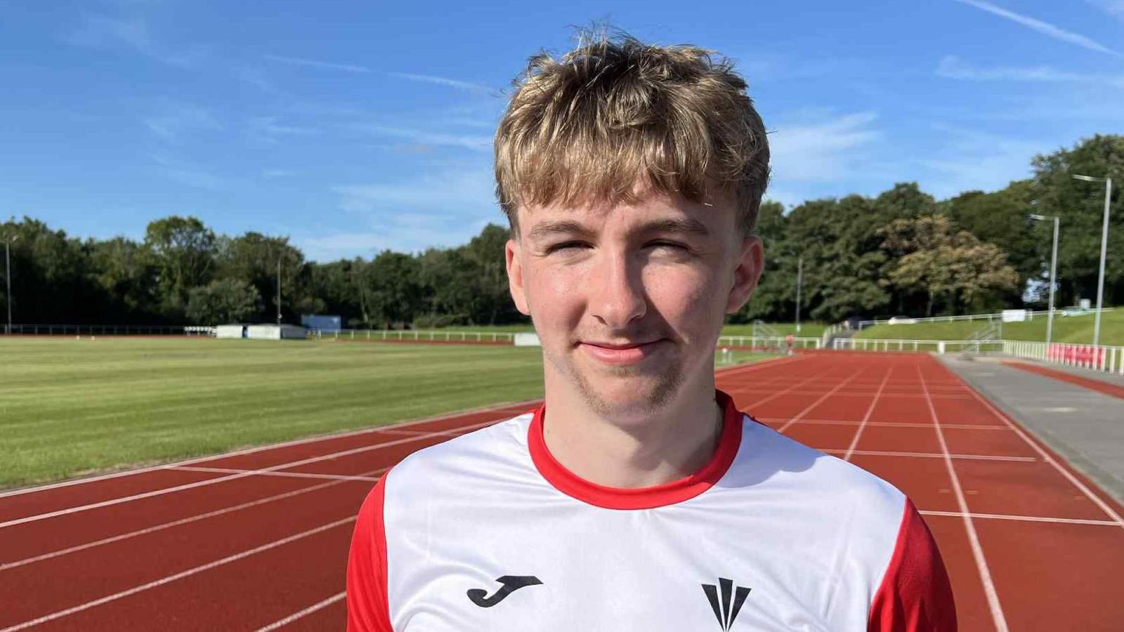 Close up picture of Ewan smiling looking at camera. His eyes are squinting due to the sun. Behind him is an athletics running track with trees in the background. Ewan wearing a white and red top and has blonde hair.