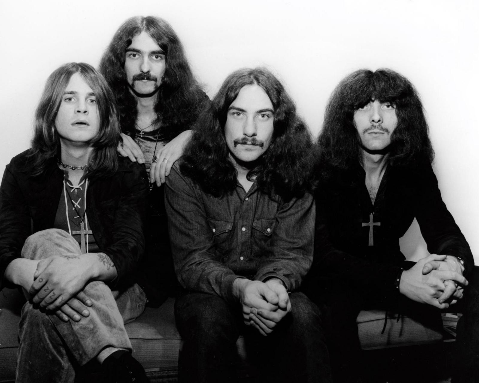 A black and white photograph shows a group of four men sitting together against a plain white backdrop. They all have long hair.