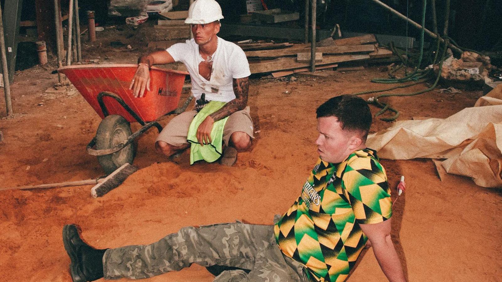 Two men lounging around on a construction site, one of them has a white hardhat and is leaning on a wheelbarrow, the other one is in a green, black and yellow Arsenal top
