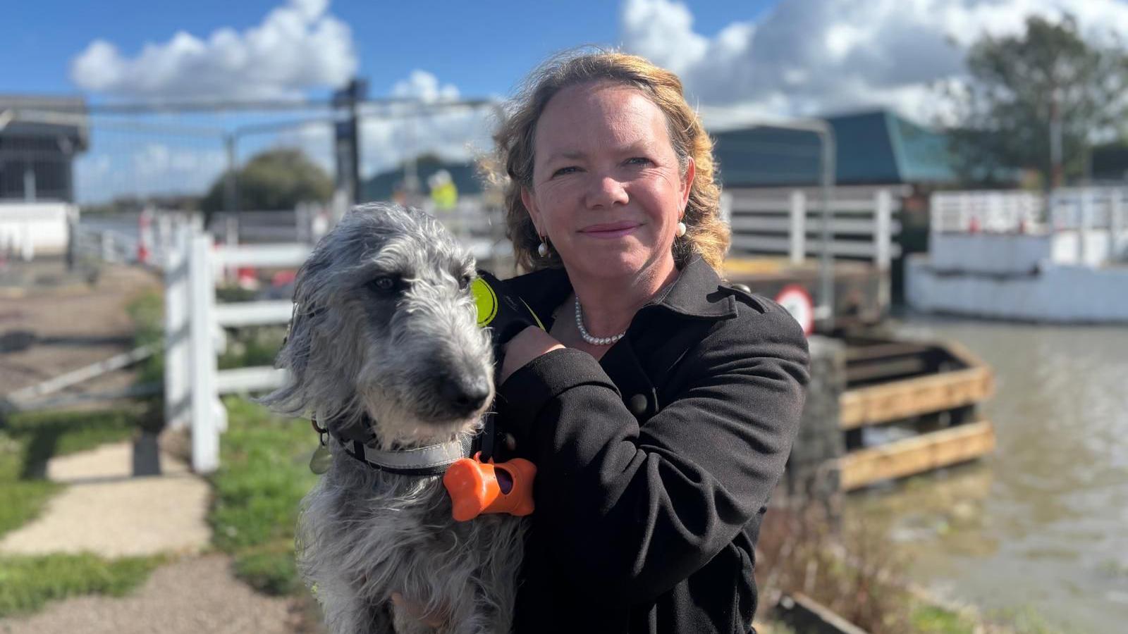 A woman in a black jacket, smiling at the camera, holds a grey dog. Behind her are the canal and Netheridge Swing Bridge.