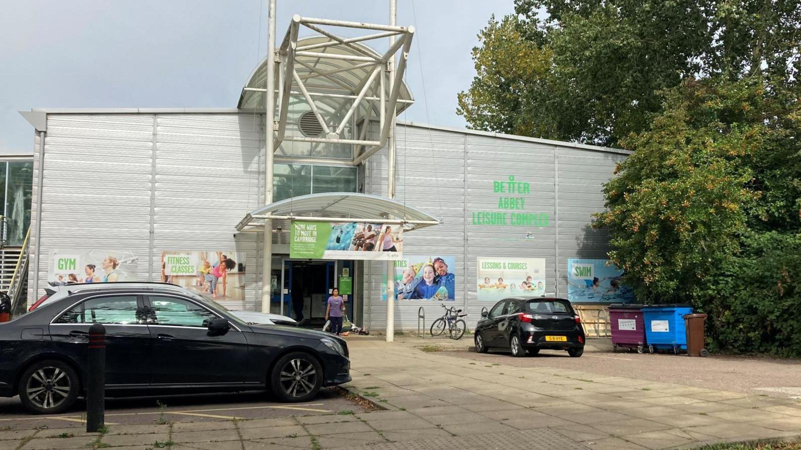 Exterior of Abbey Leisure Complex. The building is a silver-coloured warehouse-type building. Cars are parked and there's a bicycle outside. A woman is leaving the building.