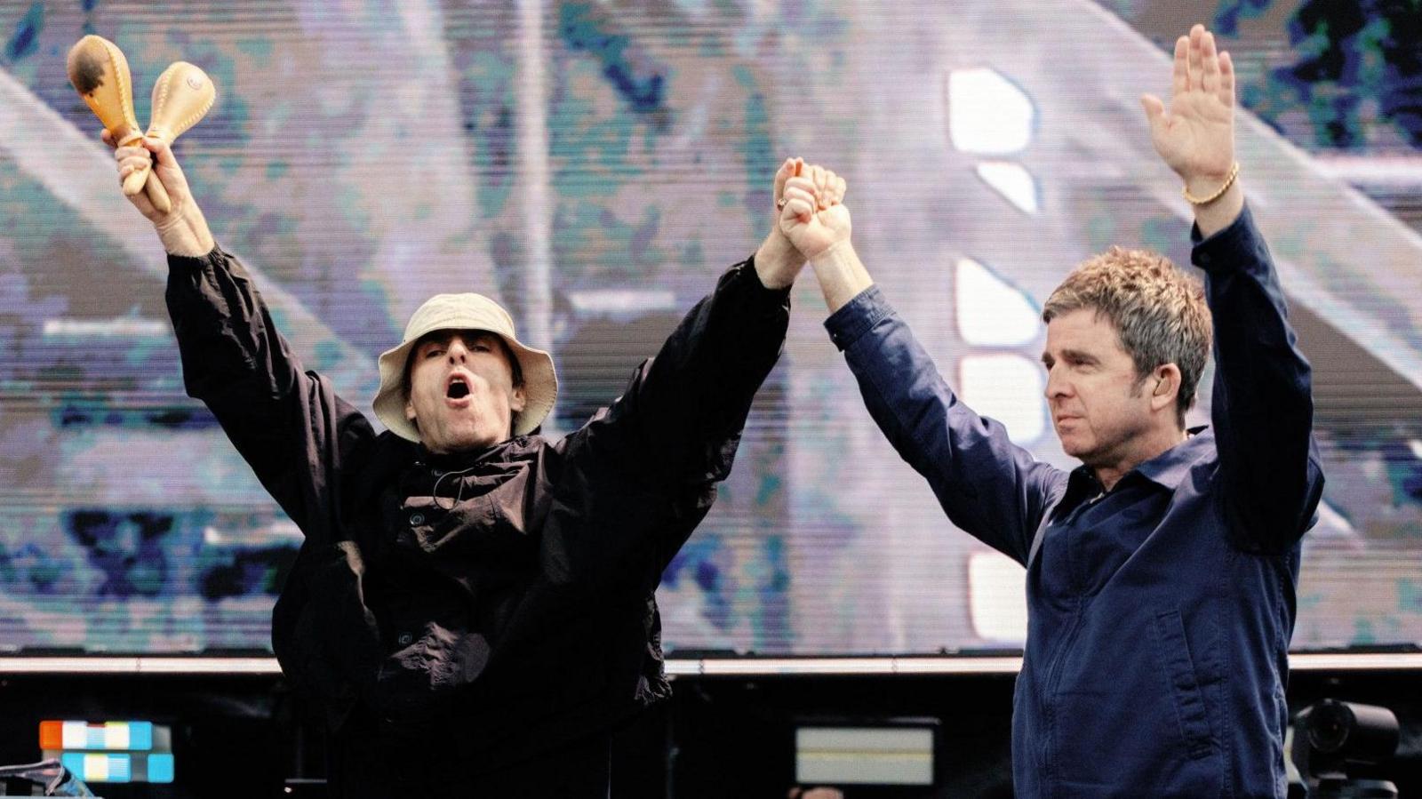 Liam and Noel Gallagher with their hands in the air as they walk on stage. Liam is wearing a beige bucket hat and a brown jacket and holding a pair of wooden maracas in the right hand. Noel is wearing a blue blazer-style jacket.