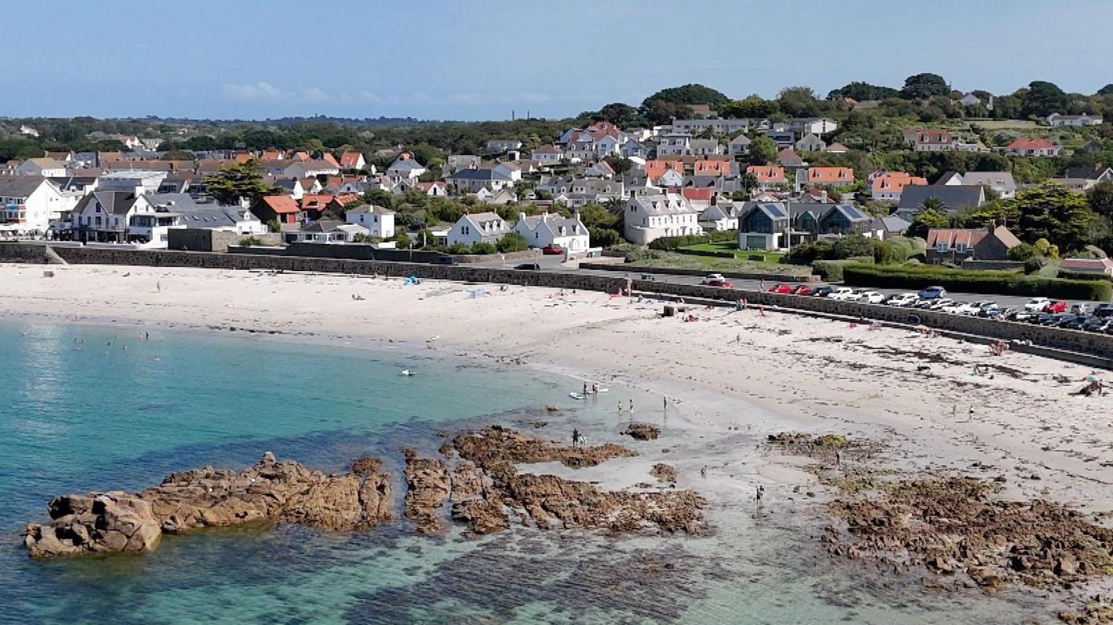 Cobo Bay in Guernsey during the summer. White sand stretches along the coast road. people are in the turquoise water. A rock formation in the sea is surrounded by seaweed. Behind the road are white houses with terracotta roofs. 