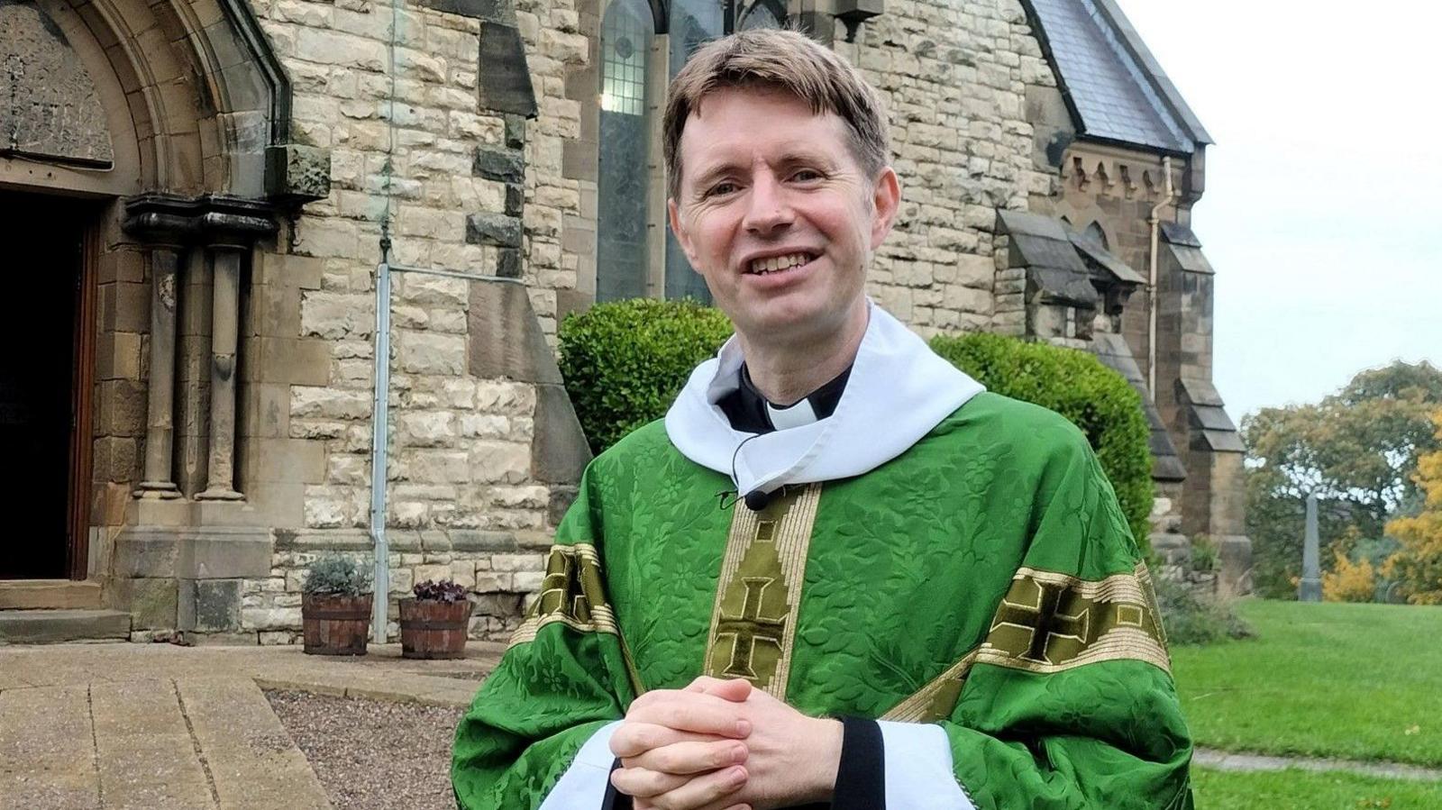 David Chadwick is wearing a green cassock is standing outside his church. He is in his 40s, clean-shaven and has short brown hair with a centre parting. His hands are clasped together in front of him and he is smiling. The church is a large, stone construction with high arch stained glass windows and an ornate arch at the entrance.