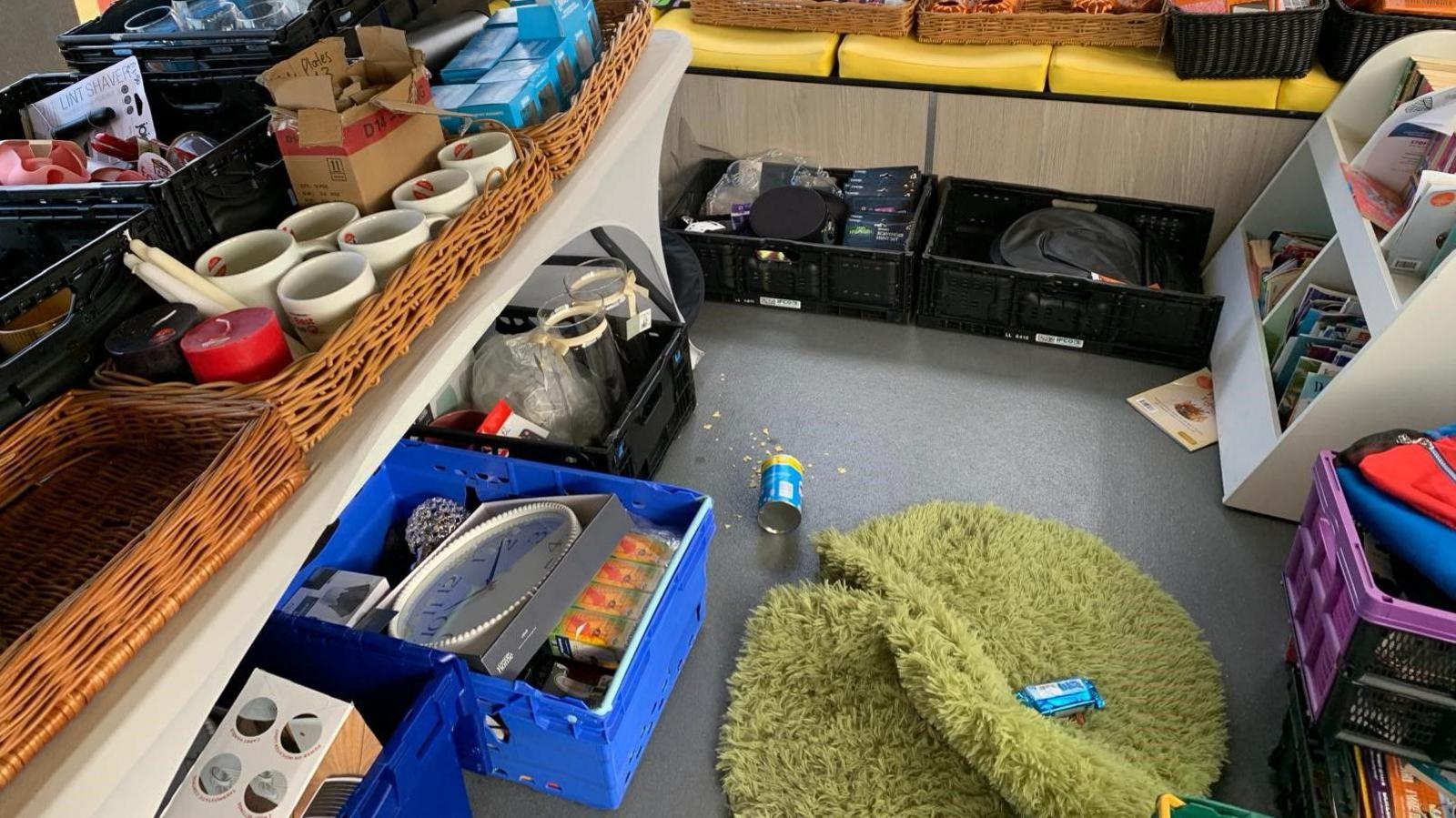 The inside of the bus which is covered in boxes of miscellaneous stuff. There is a wall clock in one of the boxes on the floor along with what looks like vases. On the countertops there are wooden boxes with candles. There is a shaggy green rug on the floor which has been folded slightly with a blue Squares bar on top of it. An open blue Pringles tube is next to the rug with some of the crisps broken around it.