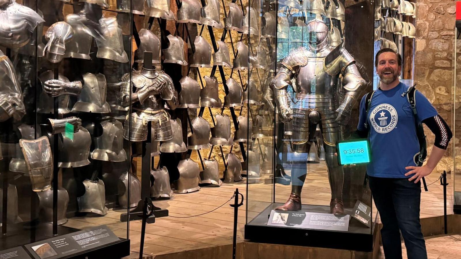 Ben Melham in a museum with lots of suits of armour. He is looking pleased and is holding a tablet with the timing on, as proof he has made the record.