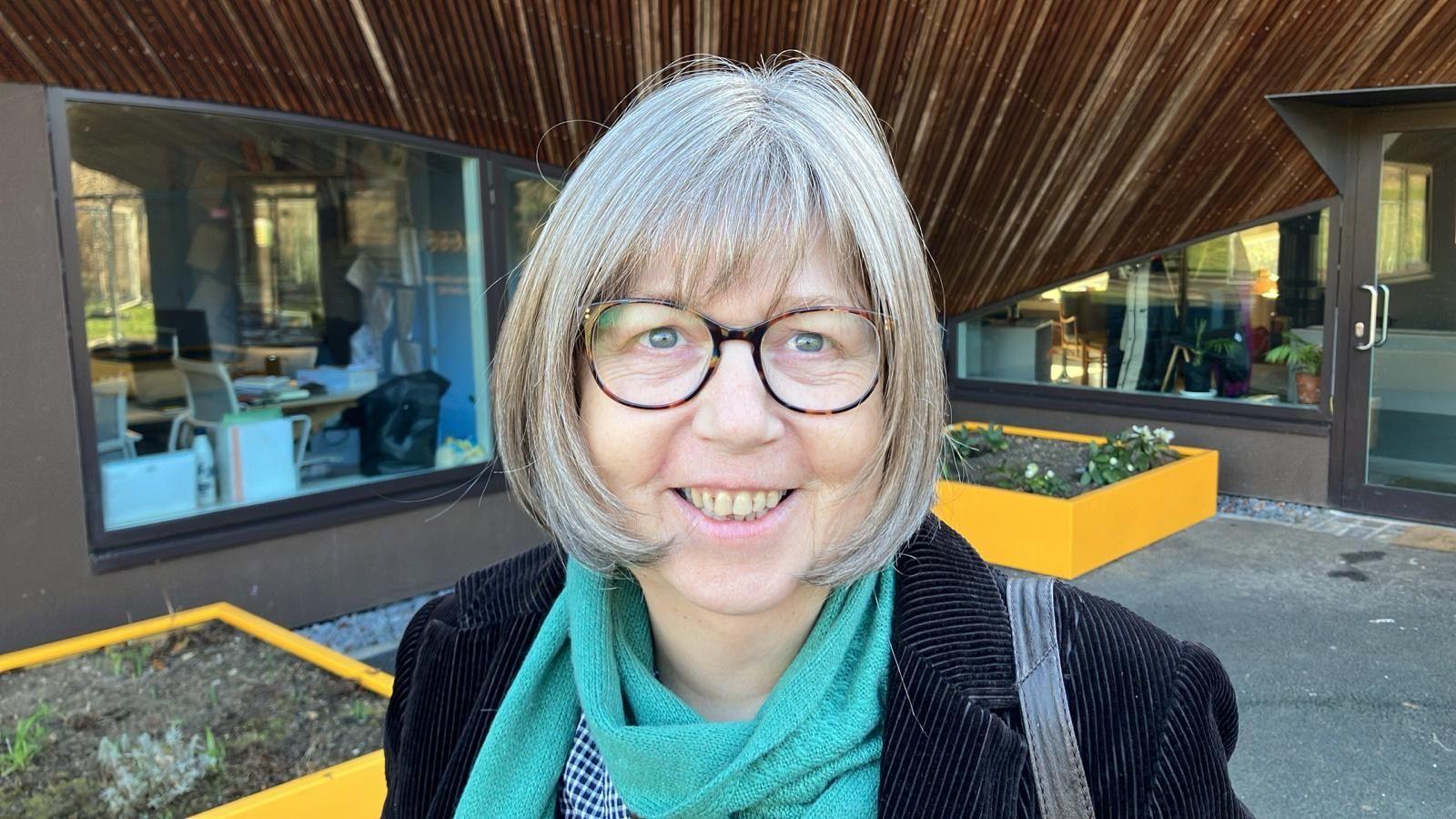 Tè le speuclairean, is falt meadhanach-ghoirid liath, le gàire air a h-aodann, na seasamh ann an gàrradh togalach phoblach 