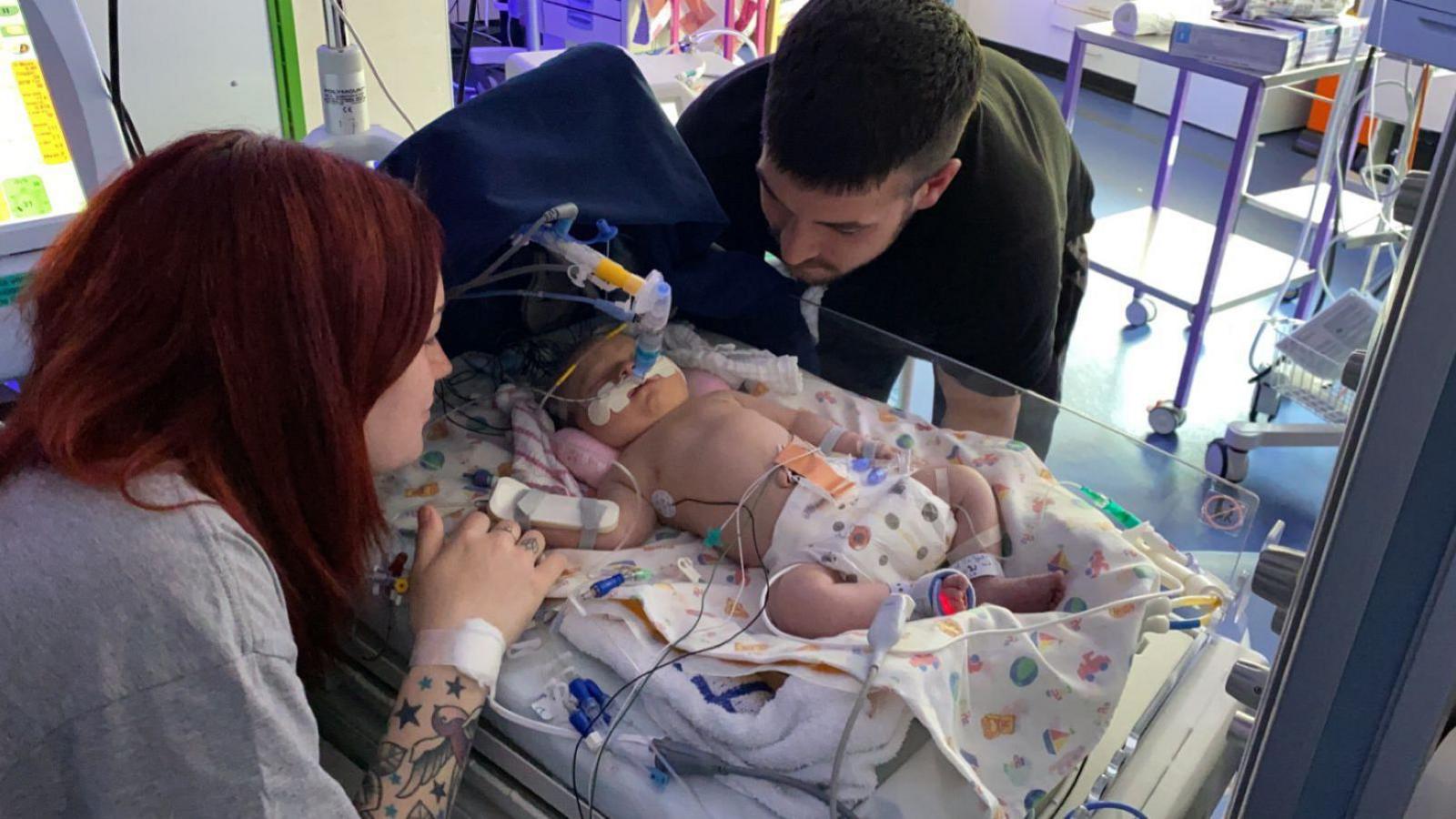 Tia with dark red hair sits beside a hospital cot, her tattooed arm reaching towards the baby in the middle. Arabella is wearing a nappy, on a ventilator and has wires and tubes attached to her. Blaze is stood being over looking down at her on the other side of the cot. Medical trolleys and paraphernalia visible in the background.