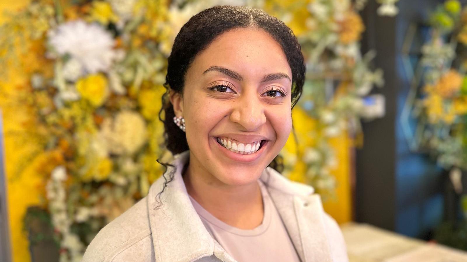 A woman with tied-back black hair wearing an earring in her right ear, a light-purple shirt and a white jacket, is smiling while standing in front of a flower-covered wall in a restaurant.