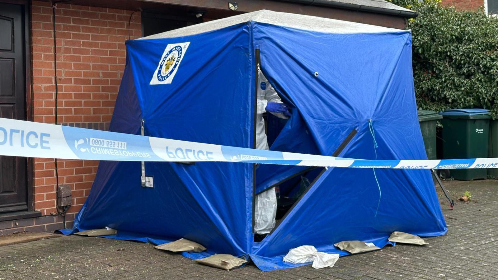 A blue tent outside a residential property. A forensics officer is inside. He is wearing a white suit and blue gloves. There are some bins nearby and police tape can also be seen. 