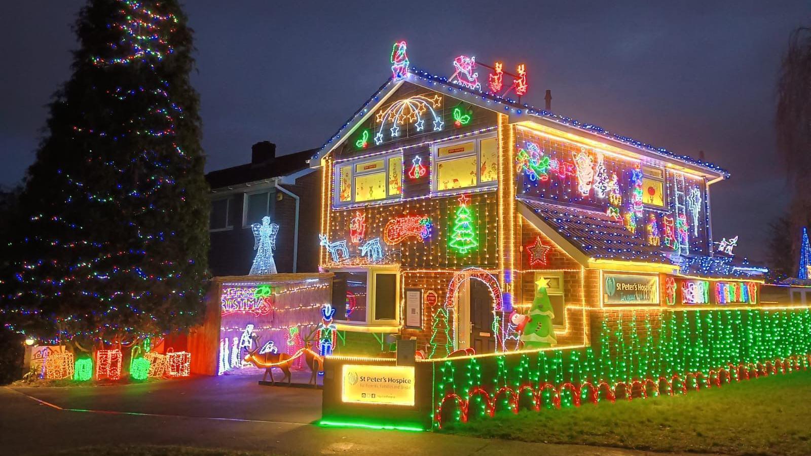 A house in Patchway, Bristol, decorated with hundreds of Christmas lights. There is a large Christmas tree outside, covered in multicoloured fairy lights. The house walls are decorated with green and red lights. The house has stars, trees, angels and Santa in a sleigh on it, all made out of lights