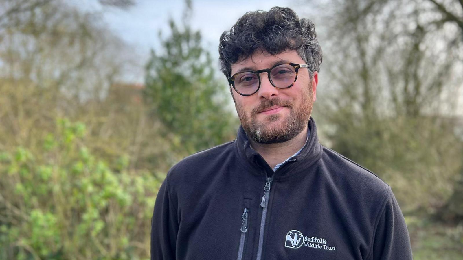 Jack Cripps is wearing glasses and a black zip-up fleece with the Suffolk Wildlife Trust logo. He is standing in a woodland and looking into the camera. 