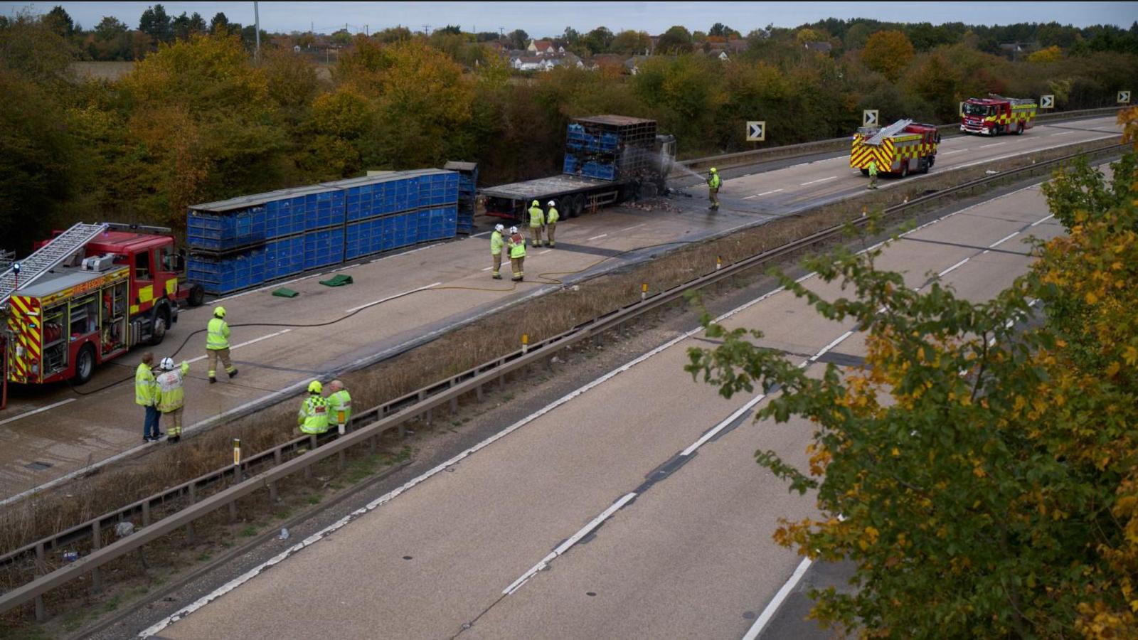 Fire engines and emergency workers on the A12. The picture is taken from above, from a bridge. A large blue container has been removed from a truck and a firefighter is hosing the cab down