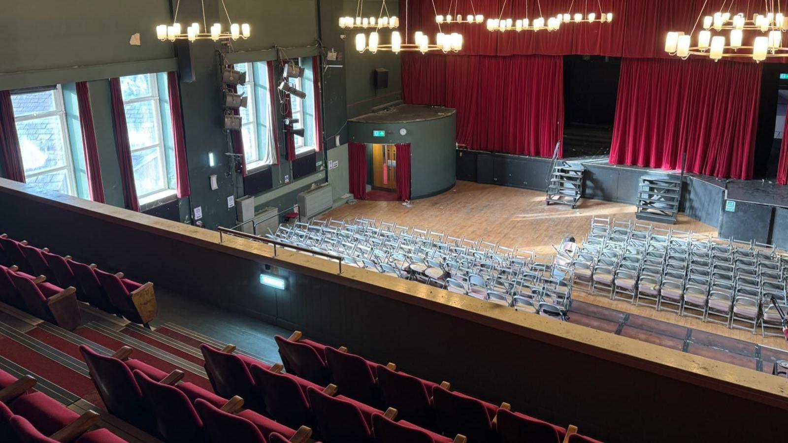 View from the balcony of the empty Oswaldtwistle Civic Arts Centre and Theatre showing red seats and on the ground level dozens of grey foldable chairs with red curtains slightly open on the stage.