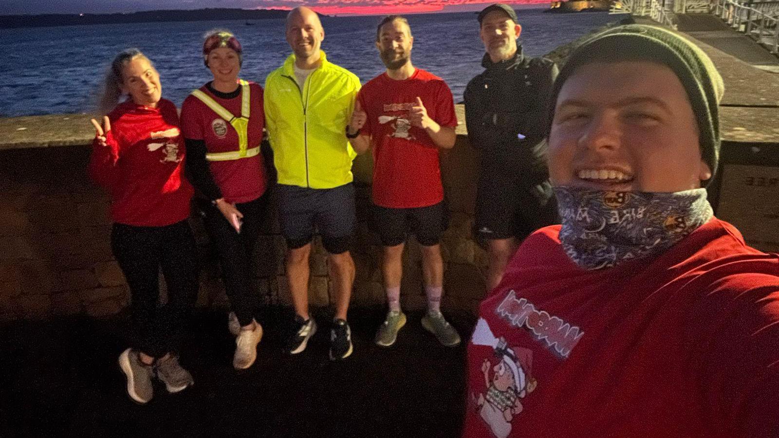 A group of runners pose for a selfie at night as the sun sets creating a red skyline over the sea behind them. They are smiling and one runner is giving a thumbs up sign to the camera and another is giving a peace sign.