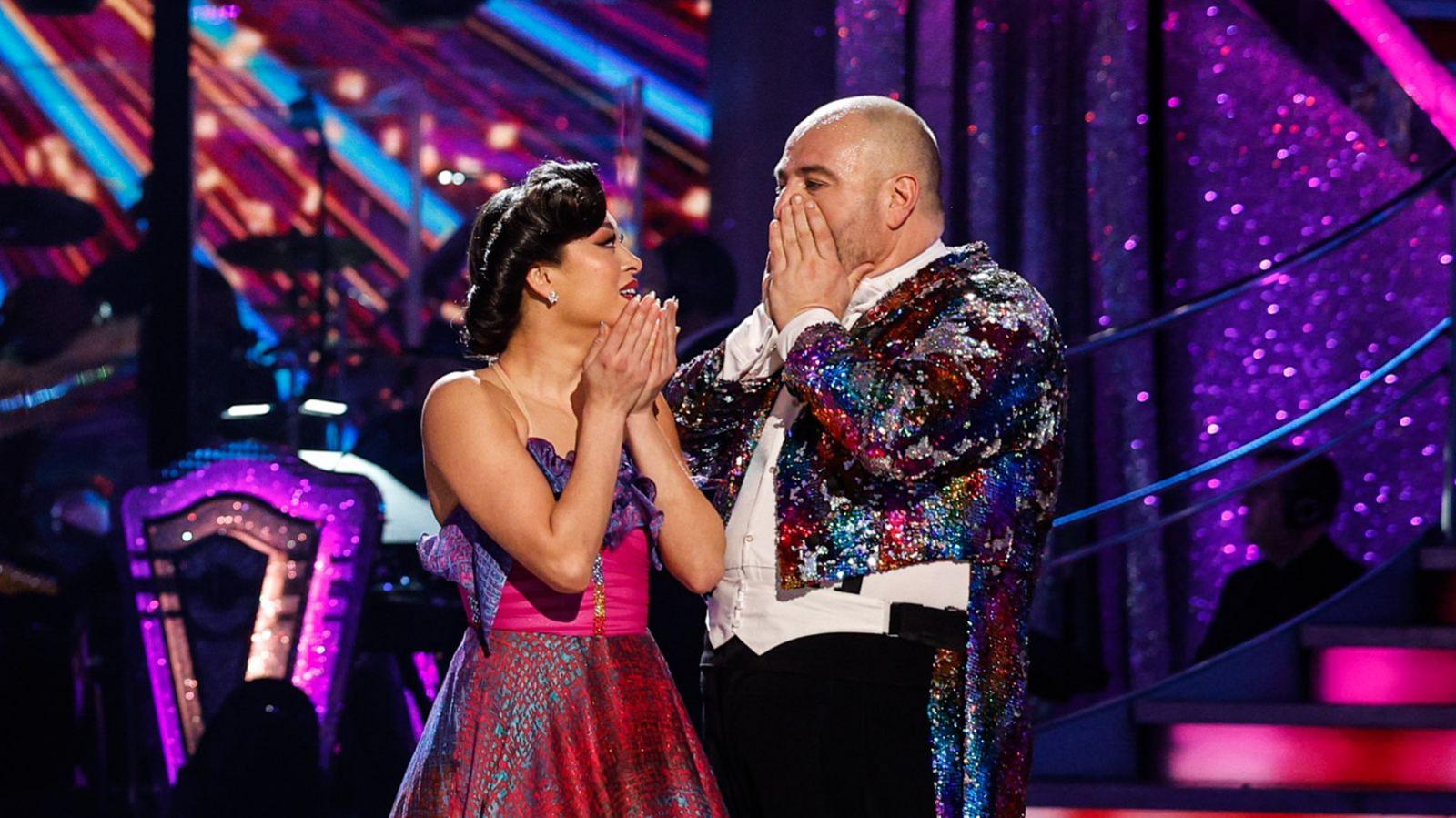 Katya Jones and Wynne Evans on the Strictly Dance Floor. Katya is on the left and is cupping her hands in front of her face. She is wearing a pink and purple desk. Wynne is to her right and has his hands covering his mouth, he is wearing black trousers and a white shirt. He is also wearing a sparkly circus masters jacket.