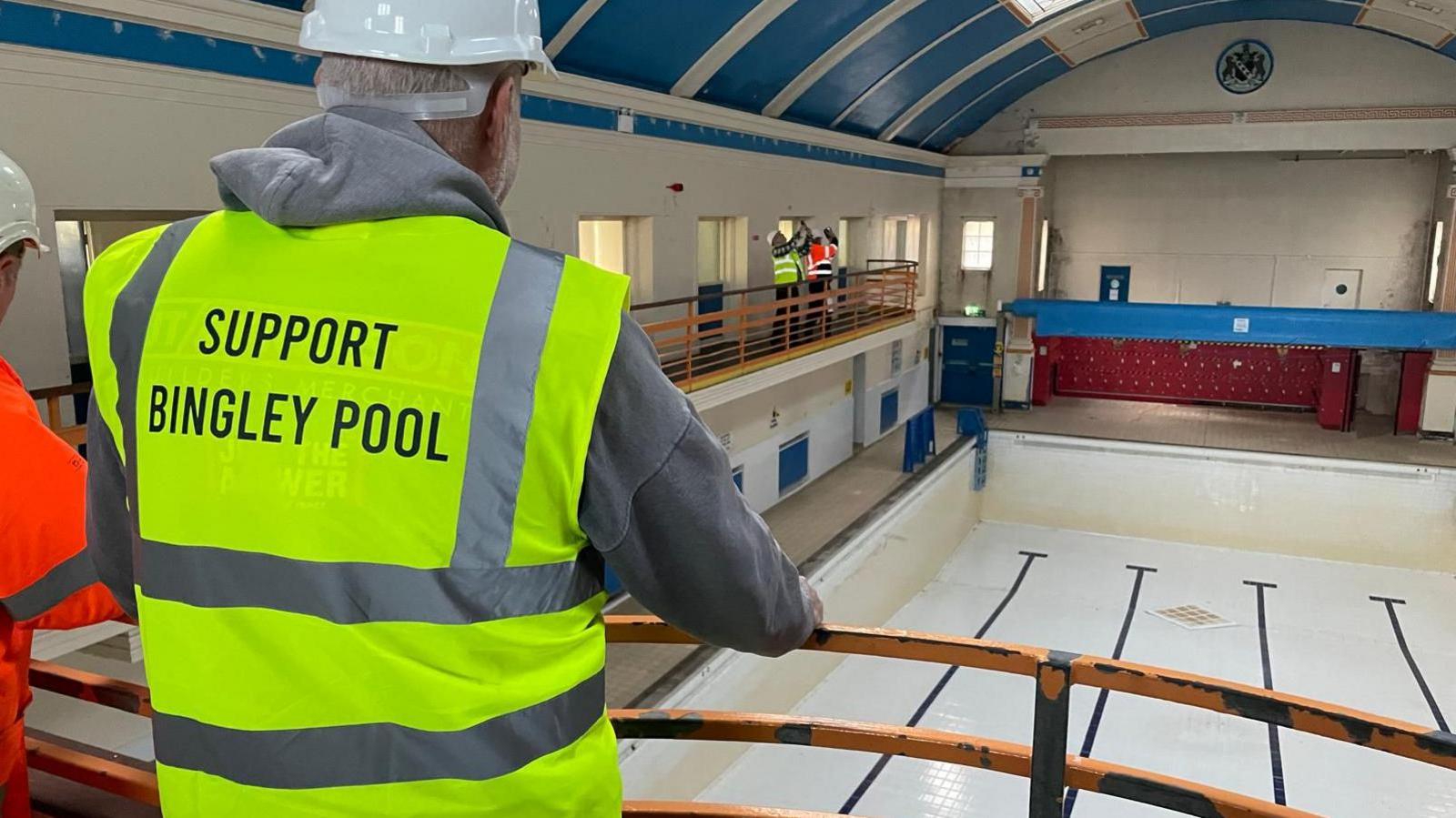 The back of a man's head with a white protective helmet and a yellow tabard looking over an empty indoor pool