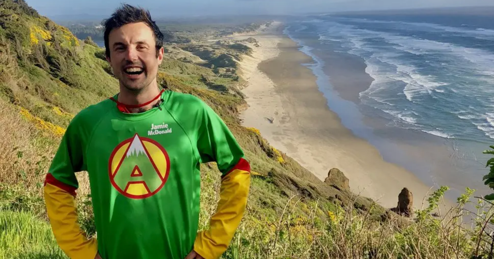 Jamie McDonald stood at the top of a grassy cliff, smiling into the camera. A beach, with crashing waves, can be seen in the distance. The sun is shining.