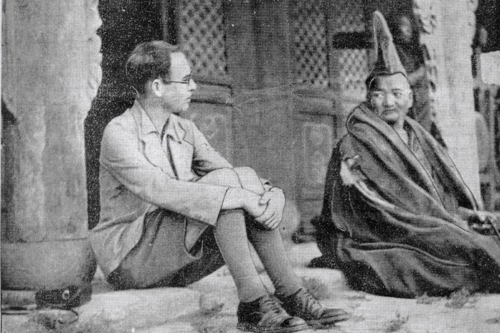 Gareth Jones sitting on the steps in front of a Buddhist temple. He is wearing dark shorts and a light jacket and is talking to a religious leader, who is to the right of him wearing a robe.