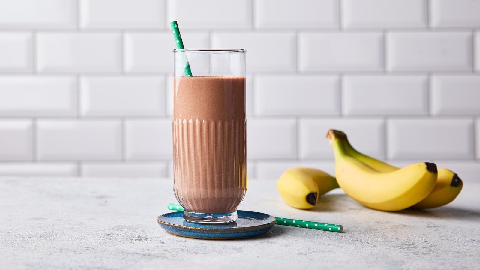 Chocolate protein milkshake in a tall glass with bananas lying on their side next to it. 