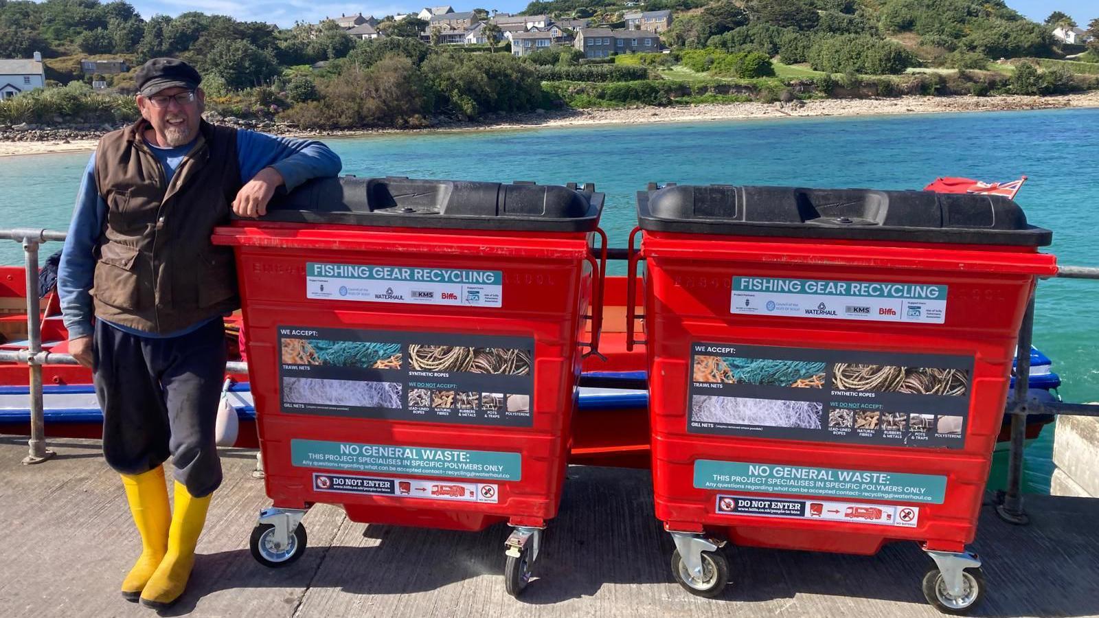 A fisherman smiling at he stands next to two large rectangular recycling bins which have labels which say: FISHING GEAR RECYCLING and NO GENERAL WASTE with pictures of fishing gear. Behind him is a bay and shoreline.