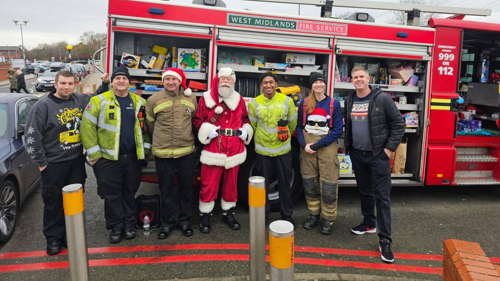 Firefighters standing alongside a fire engine, with Santa standing in the middle, wearing his traditional red costume. There are toys inside the engine behind them.