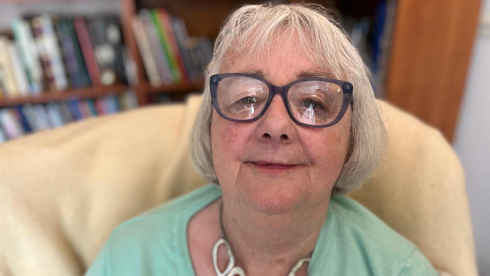 Lucille is wearing a pale green cardigan and glasses. She is sat in her living room with a bookshelf behind her.