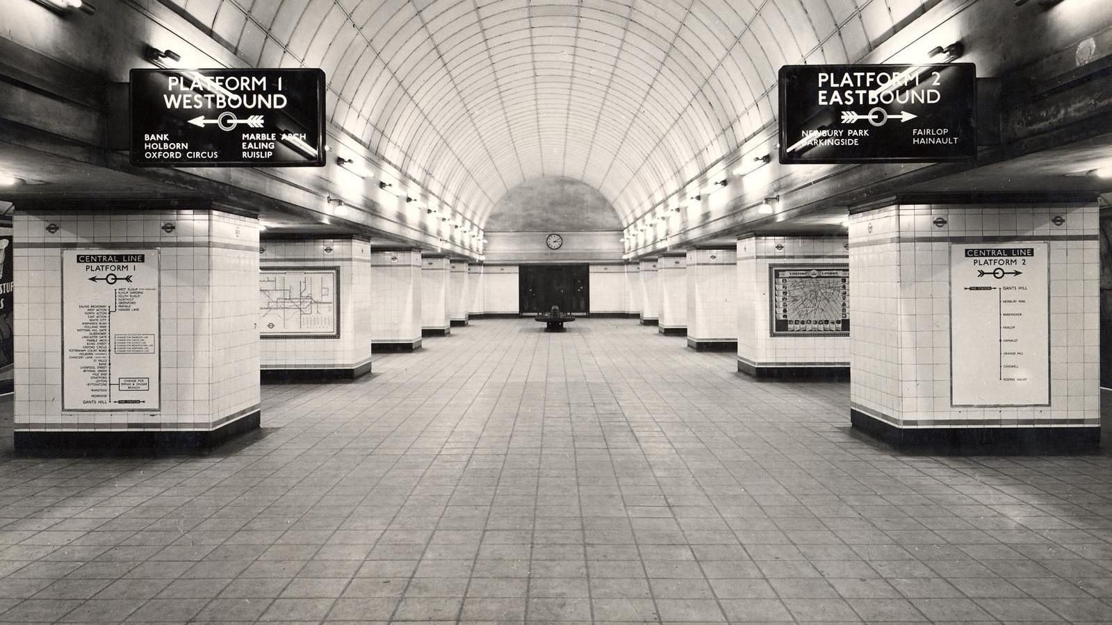 A black-and-white photo of Gants Hill tube station concourse. There is a wide, sweeping tunnel, with platforms 1 and 2 on either side. These are labelled with overhead signs, with arrows pointing to the platforms. There are also route maps on the walls to the side of each platform.