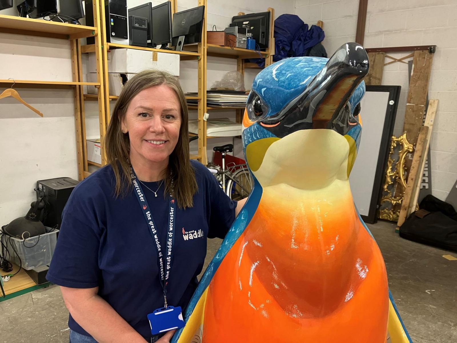 A woman wearing a blue t-shirt stands next to an orange and blue penguin sculpture