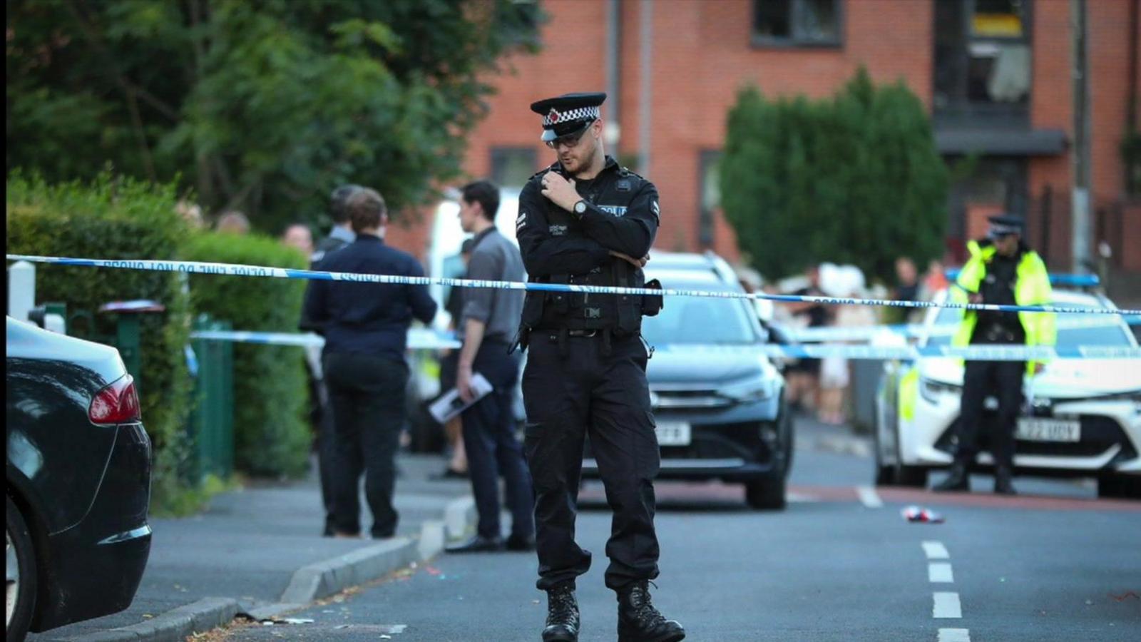 A police officer, standing behind blue and white police cordon tape speaking into to his radio on his chest. Other officers can been seen in the background next to a police car on Panfield Road