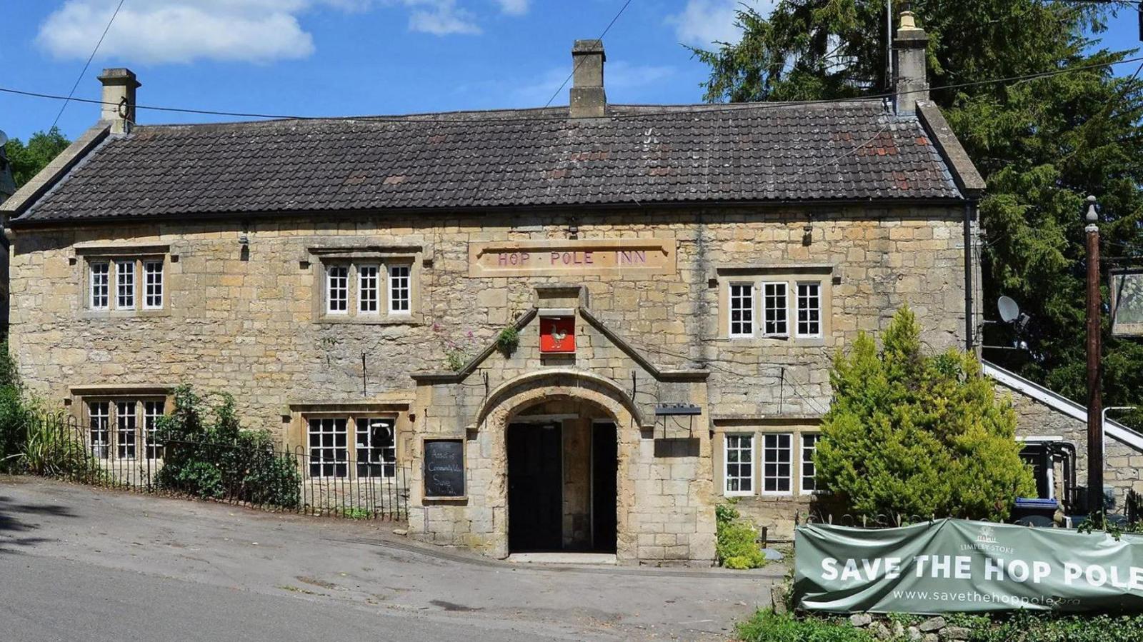 The Hop Pole pub in Limpley Stoke. It is an old two storey brick building with a pointed archway entrance and small, sunken sash windows. The roof is a dark slate and there are black metal railings separating the pub from the road, which is slanted on a hill. Outside the parking area to the right there is a green banner attached to a hedge that reads Save the Hope Pole