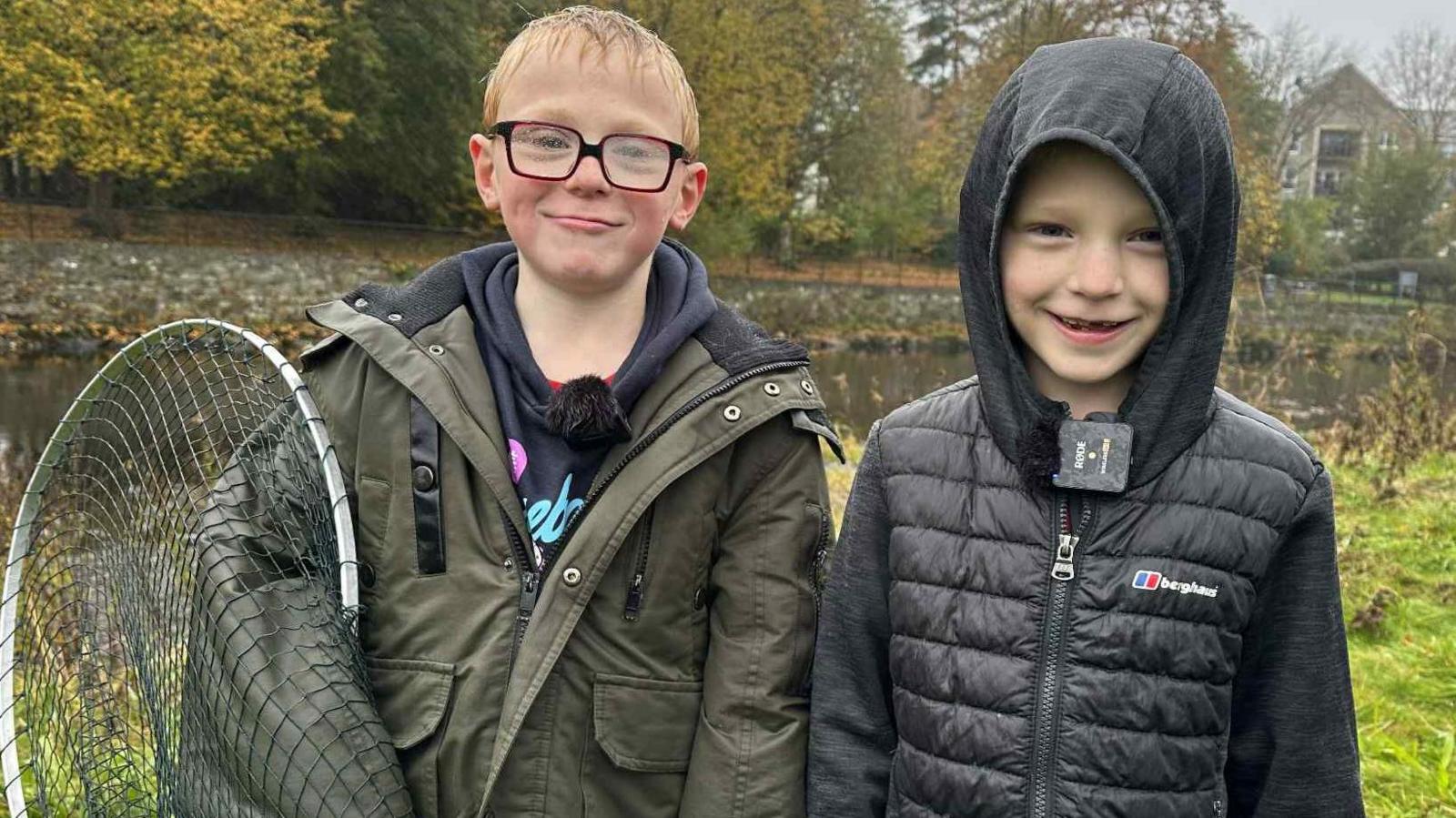 Young boys Thomas and Elijah stand together and smile at the camera. Thomas who is wearing a large green jacket and red glasses, has his right arm in a fishing net. Elijah is wearing a grey/black Berghaus puffer jacket with his hood up.