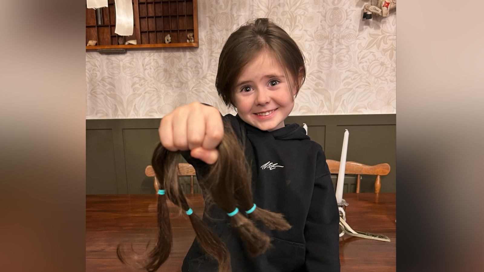 Reuben smiles at the camera while holding one arm out in front of him as he holds on to his brown hair that was cut. He wears a black hoodie as he sits on a wood table.