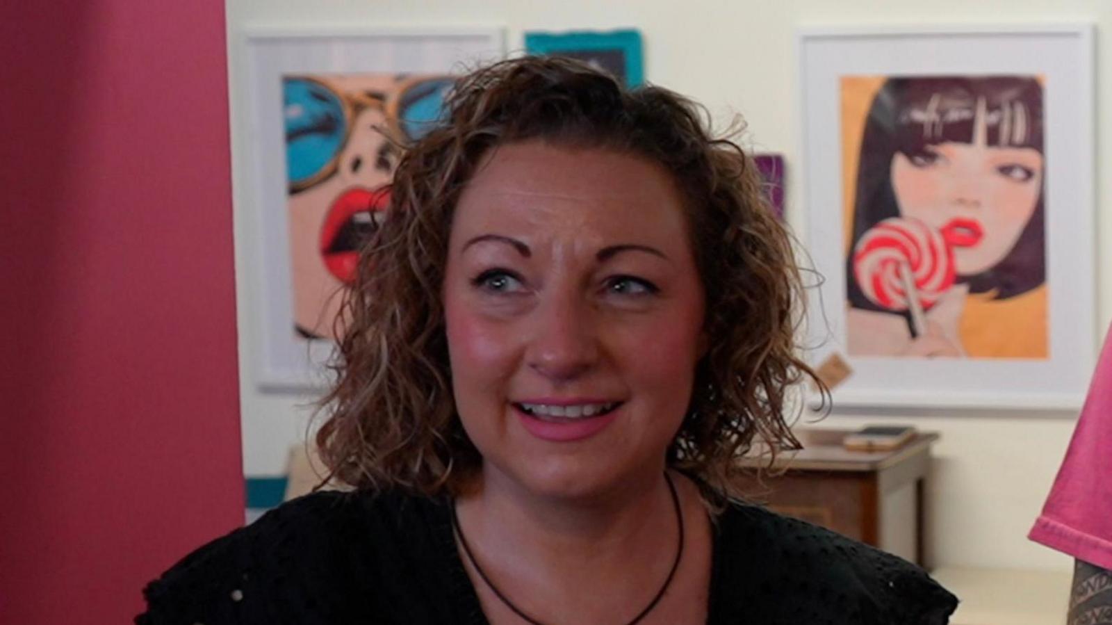 The image shows Jenny Needham standing in front of a wall decorated with multiple framed artworks. The artworks feature vibrant, retro-style illustrations, including stylized portraits, bold typography, and colourful motifs such as rainbows and candy. One prominent piece in the centre depicts a person holding a red-and-white lollipop. The frames vary in size and are arranged in a neat grid pattern, creating a visually appealing backdrop.
She wears a black short-sleeved top with a subtle textured pattern and a necklace with a small pendant.