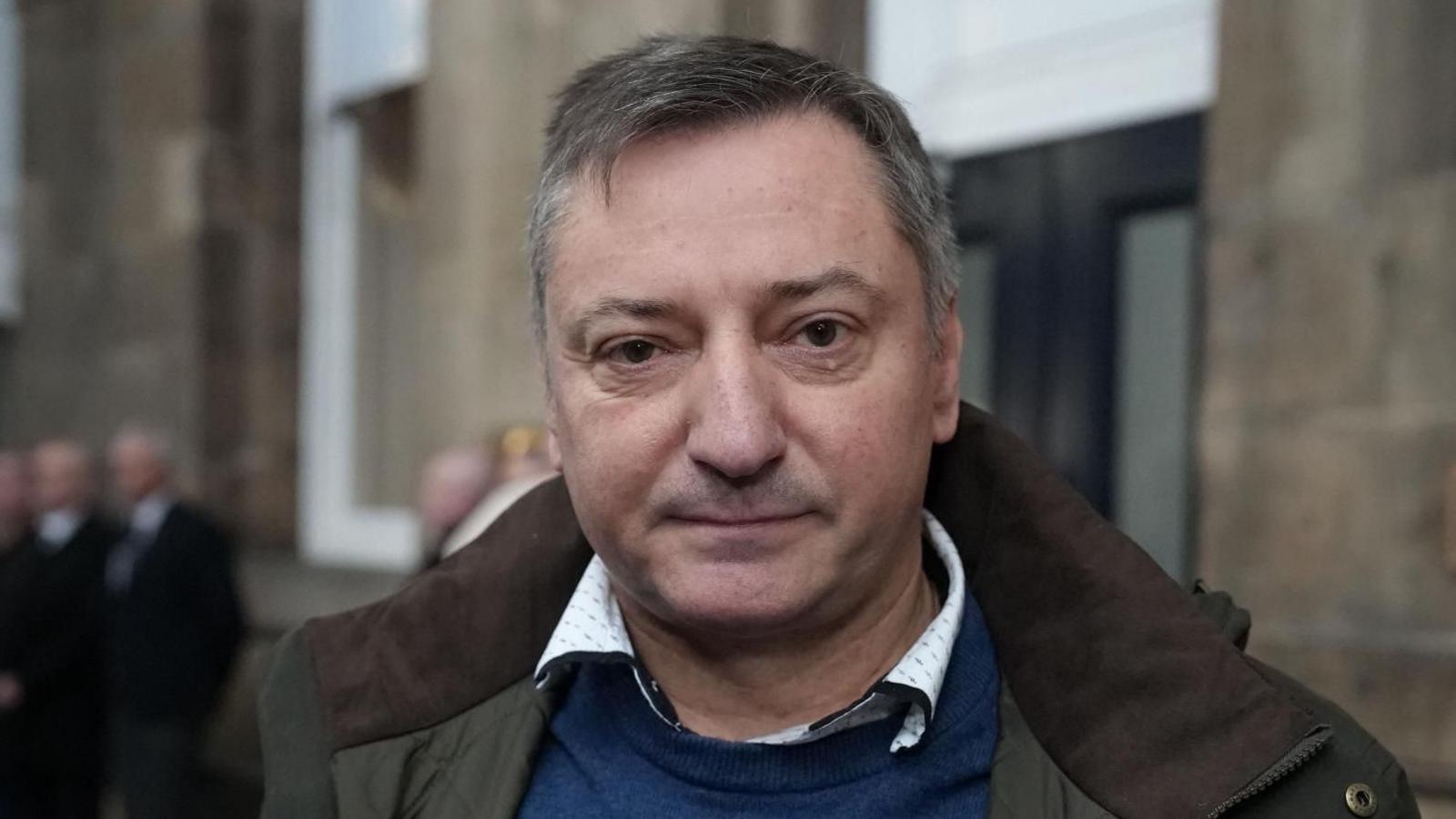 Një burrë me flokë të shkurtër gri, i veshur me një bluzë blu dhe një pallto jeshile dhe kafe, duke qëndruar në një platformë hekurudhore