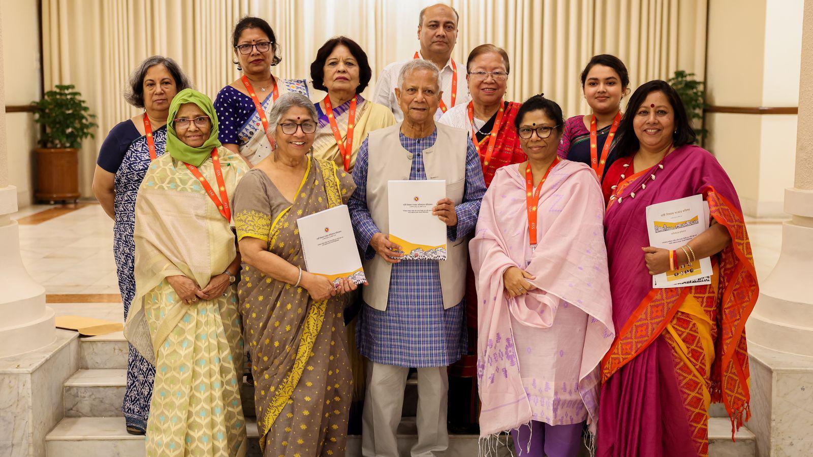 A group of 11 people stand in a marble hallway. All but two are women. The women wear bright saris, and appear to be a range of ages. In the middle is Yunus, the interim leader, wearing a blue and grey suit. He and two of the women hold reports in their hands.