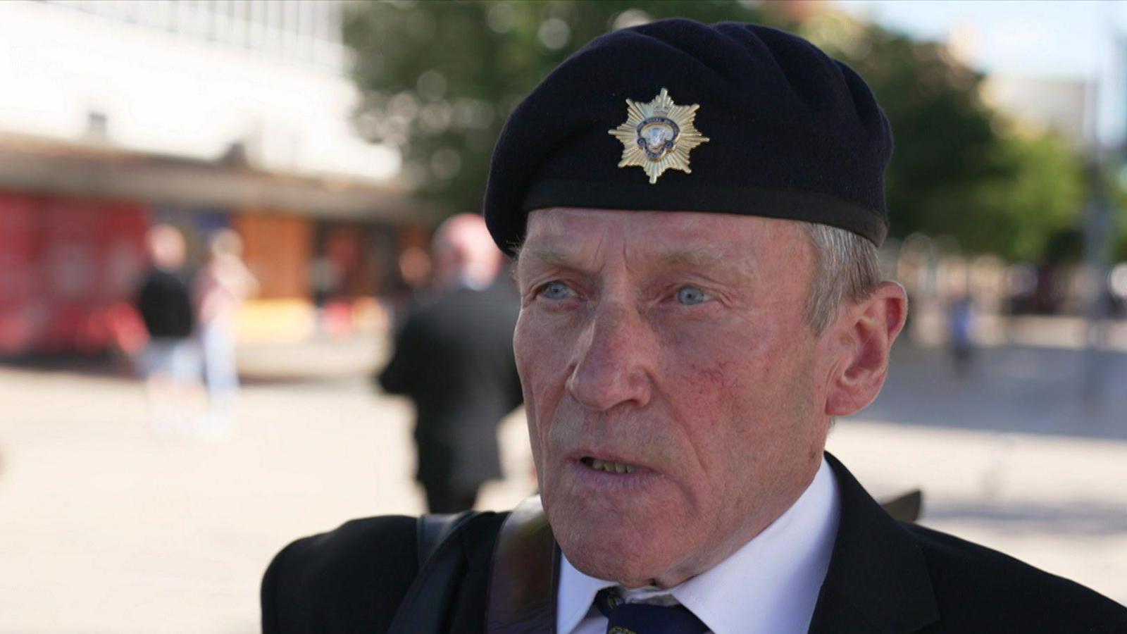 A man looking to the left of the camera, wearing a beret with a military badge on it