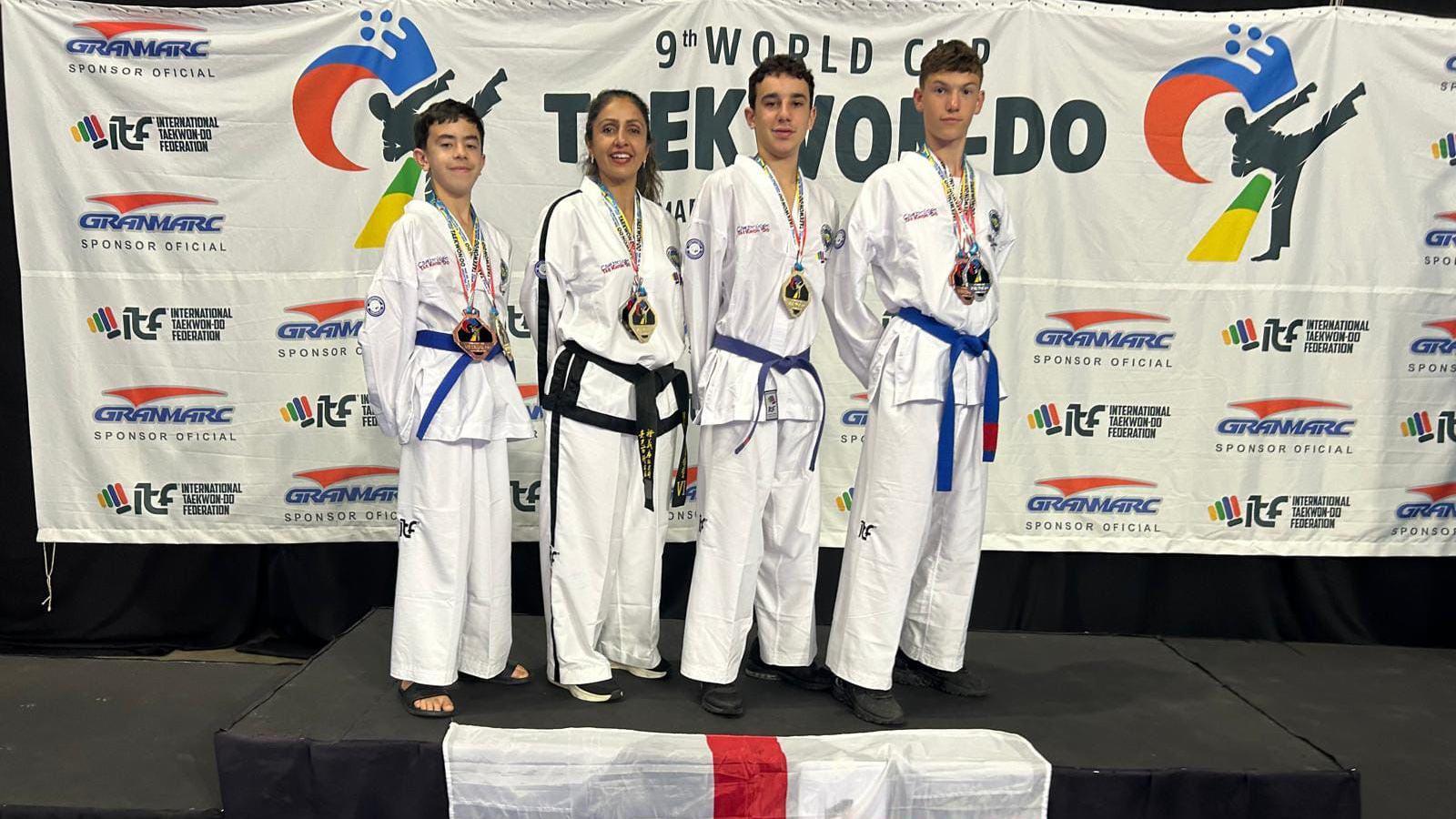 Three boys and a woman, all in white martial arts outfits, stand on a podium, upon which a St George's flag has been draped. Behind is a backdrop that says "9th World Cup Taekwon-do" and features sponsors' logos.