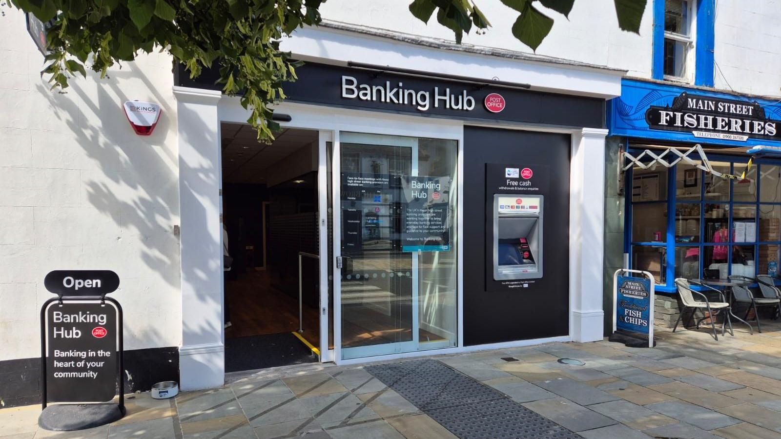 Cockermouth Banking Hub. The facility has a glass entrance door and a cashpoint. A sign for the hub is also on the pavement.