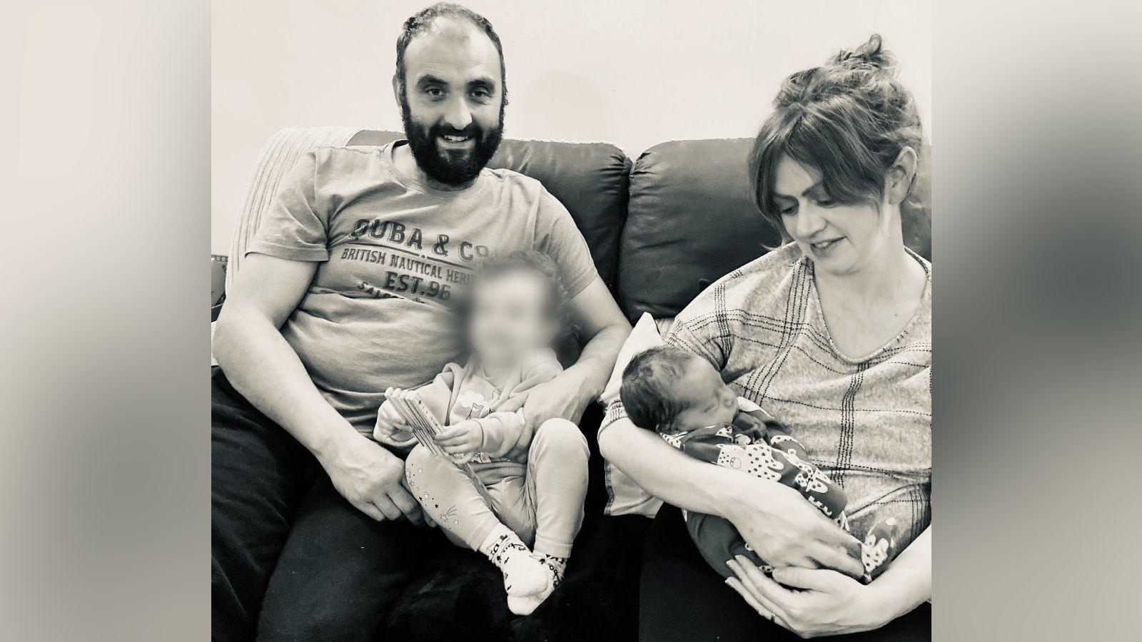 A man and a woman sitting on a sofa with two children. The man has dark hair with a receding hairline and a dark beard. He is smiling at the camera and has his arms around a toddler who has their face blurred. The woman is looking down at a sleeping baby in her arms.