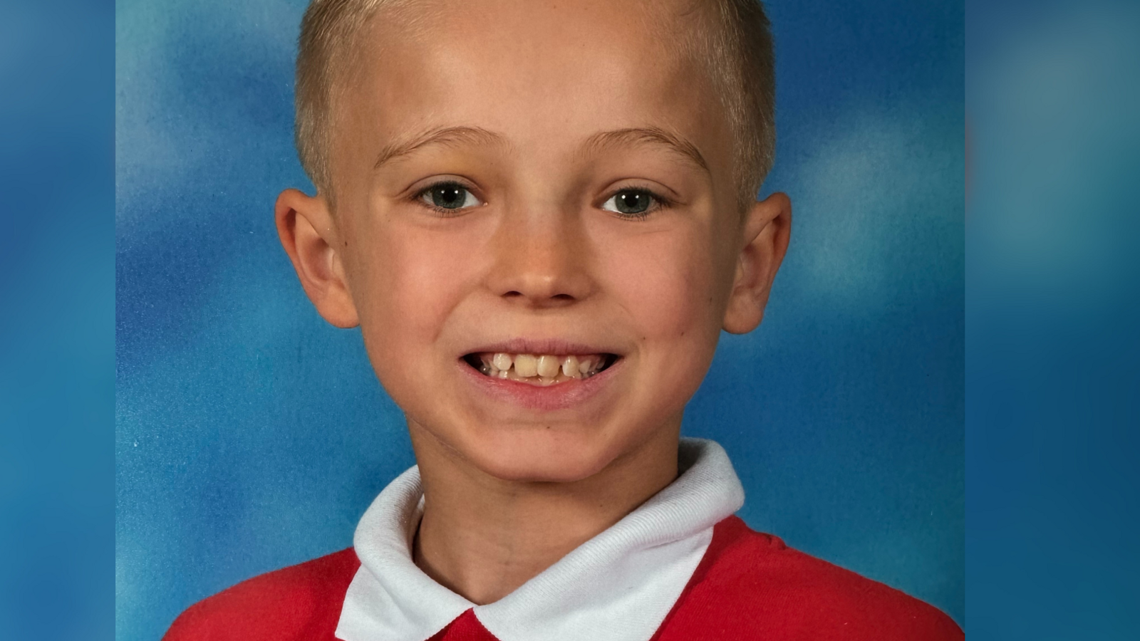 A young boy is posing in a school photo, smiling at the camera. He has blond hair and blue eyes, and is wearing a white shirt and a red jumper. The background is blue